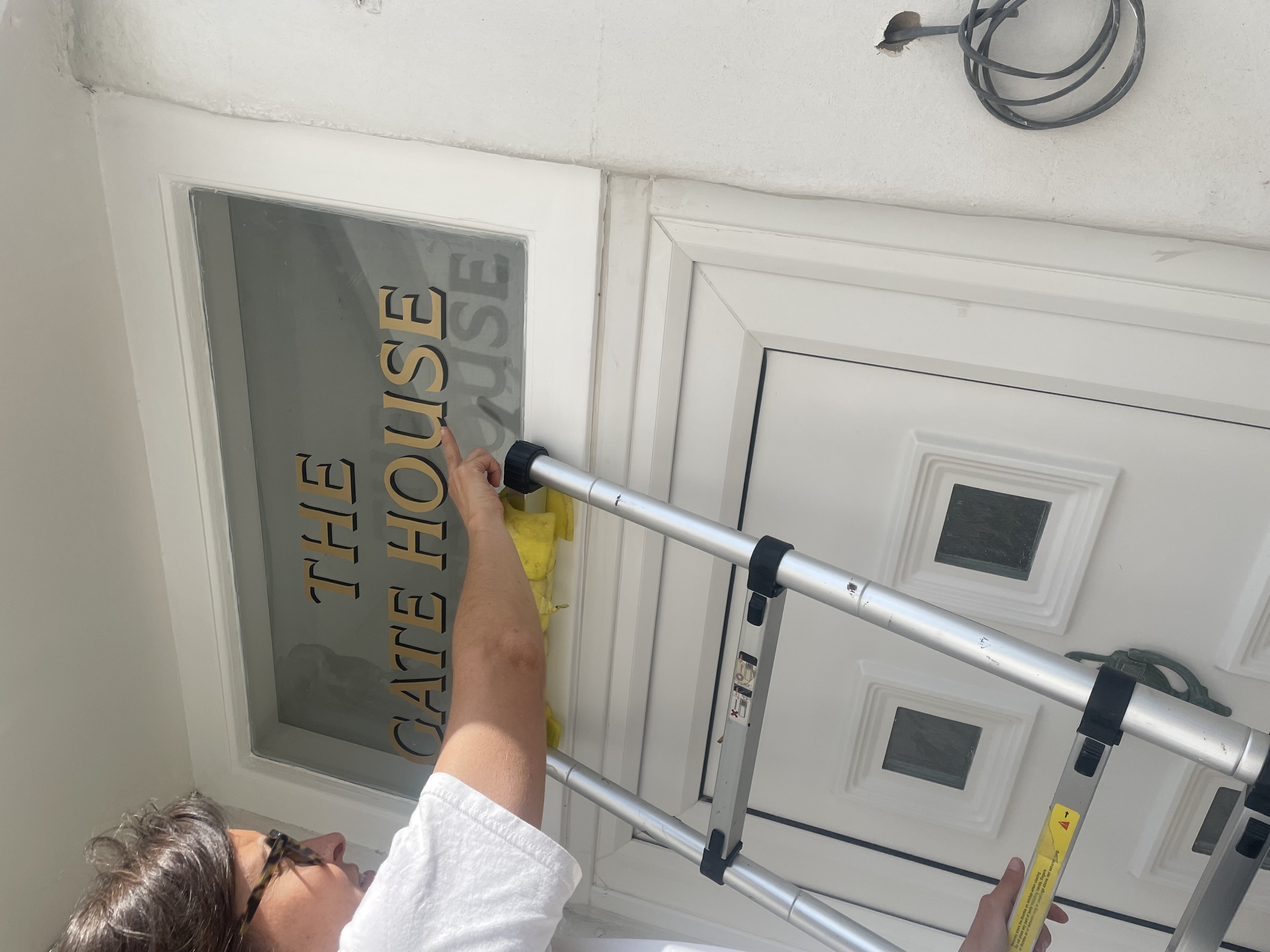 Person on a ladder pointing at the sign 'THE GATE HOUSE' White-columned Numbers sign written house entrance with gold lettering