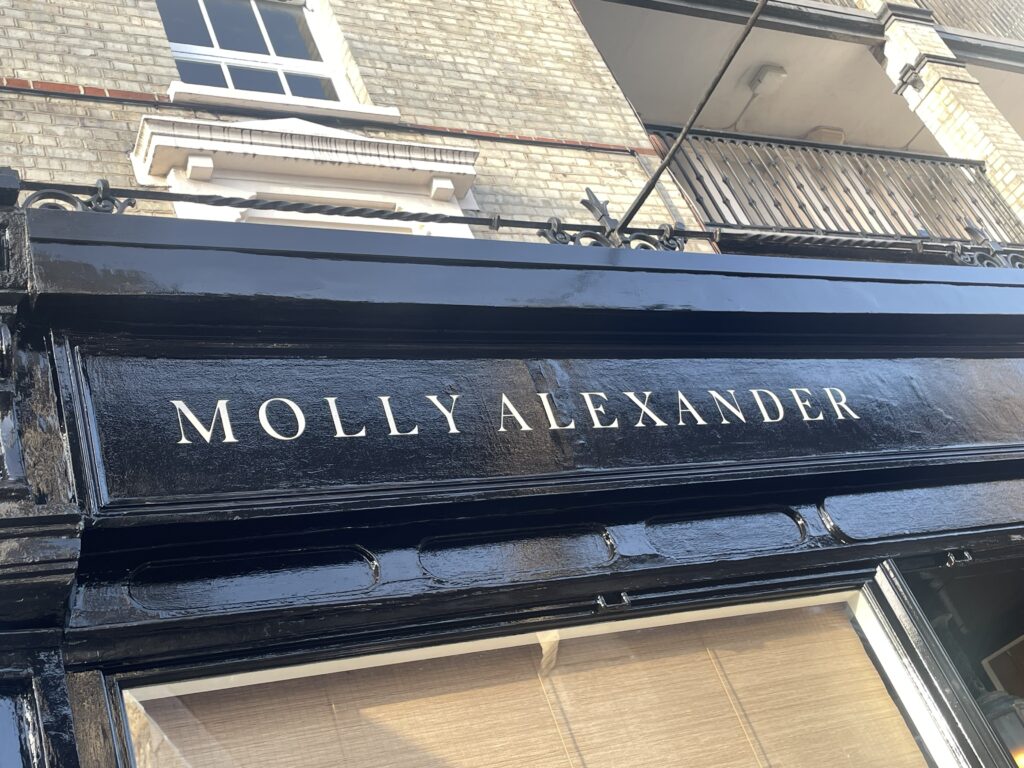 Front of a brick storefront with a glossy black sign bearing the white name 'MOLLY ALEXANDER' above the entrance.