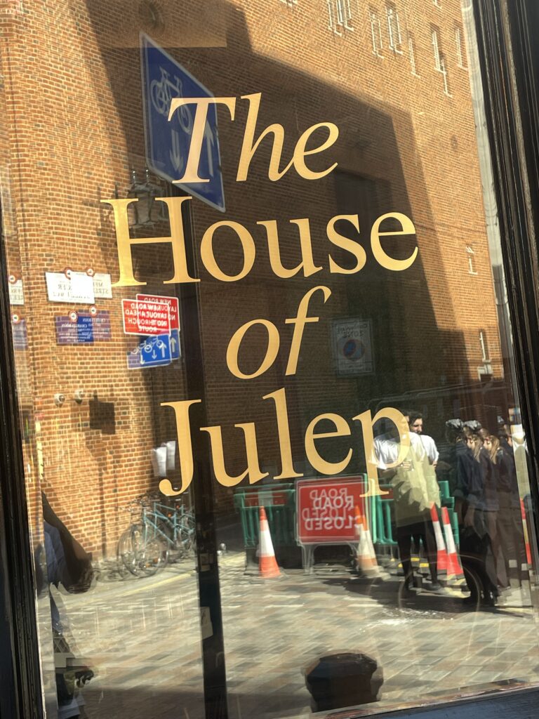 Storefront window with large gold lettering saying 'The House of' and reflections of a brick building and pedestrians outside.