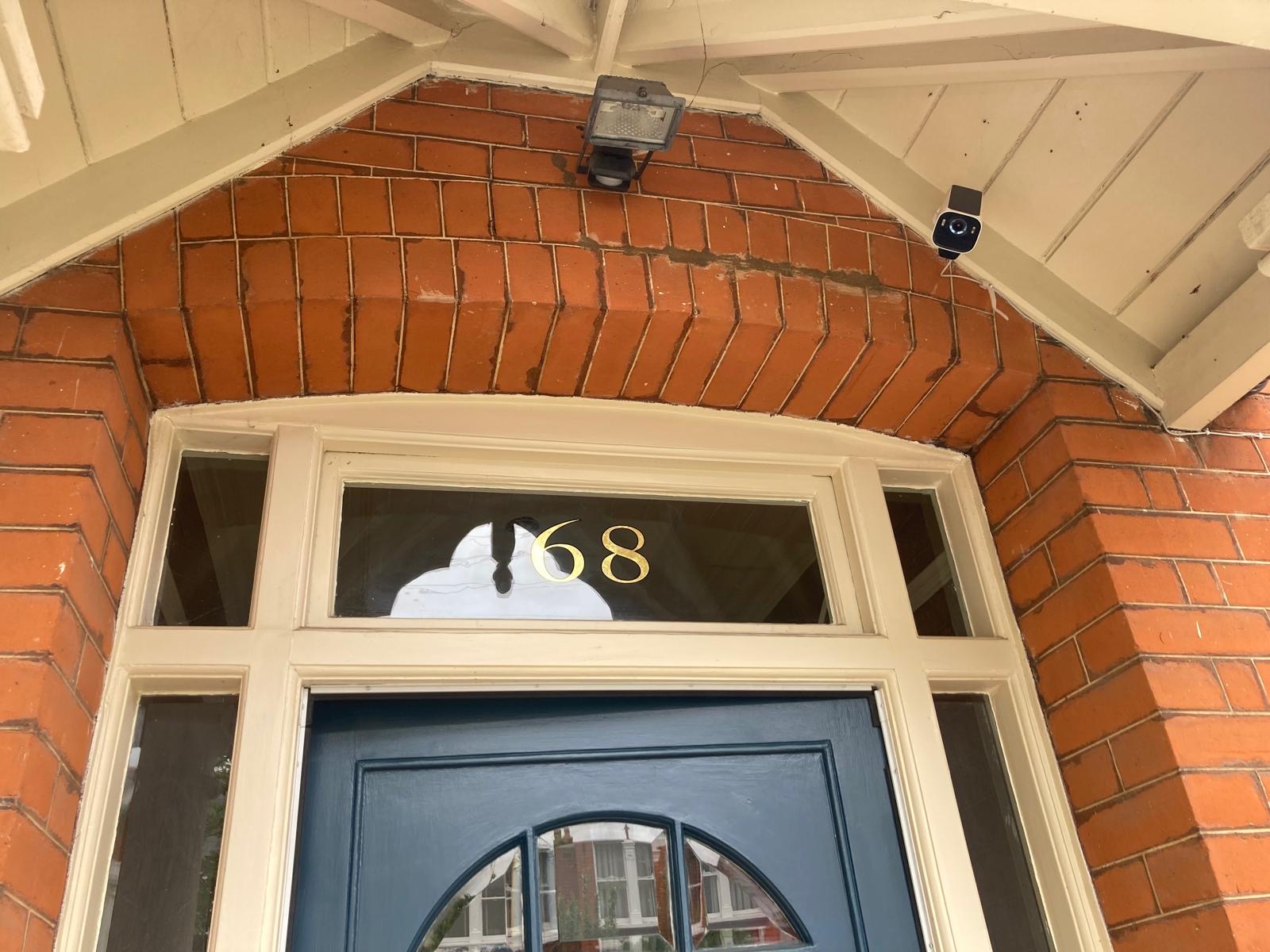 Brick entryway with a blue door under a brick arch; house number 68 visible on the transom above the door, plus a security camera and motion light under the eaves.