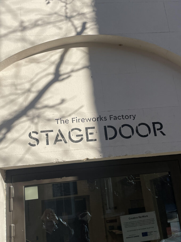 Facade of a building with a stage door sign reading 'The Fireworks Factory' and large 'STAGE DOOR' text, with tree shadows on the wall.